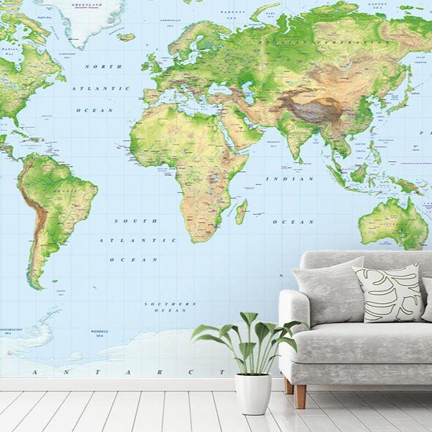 A large, detailed world map mural from Wallpapered.com featuring vibrant green landmasses, topographical shading, and labeled continents, countries, and capitals. The mural spans an entire wall behind a modern gray sofa with green-patterned cushions, complemented by a white vase with leafy greenery on a light wood floor.